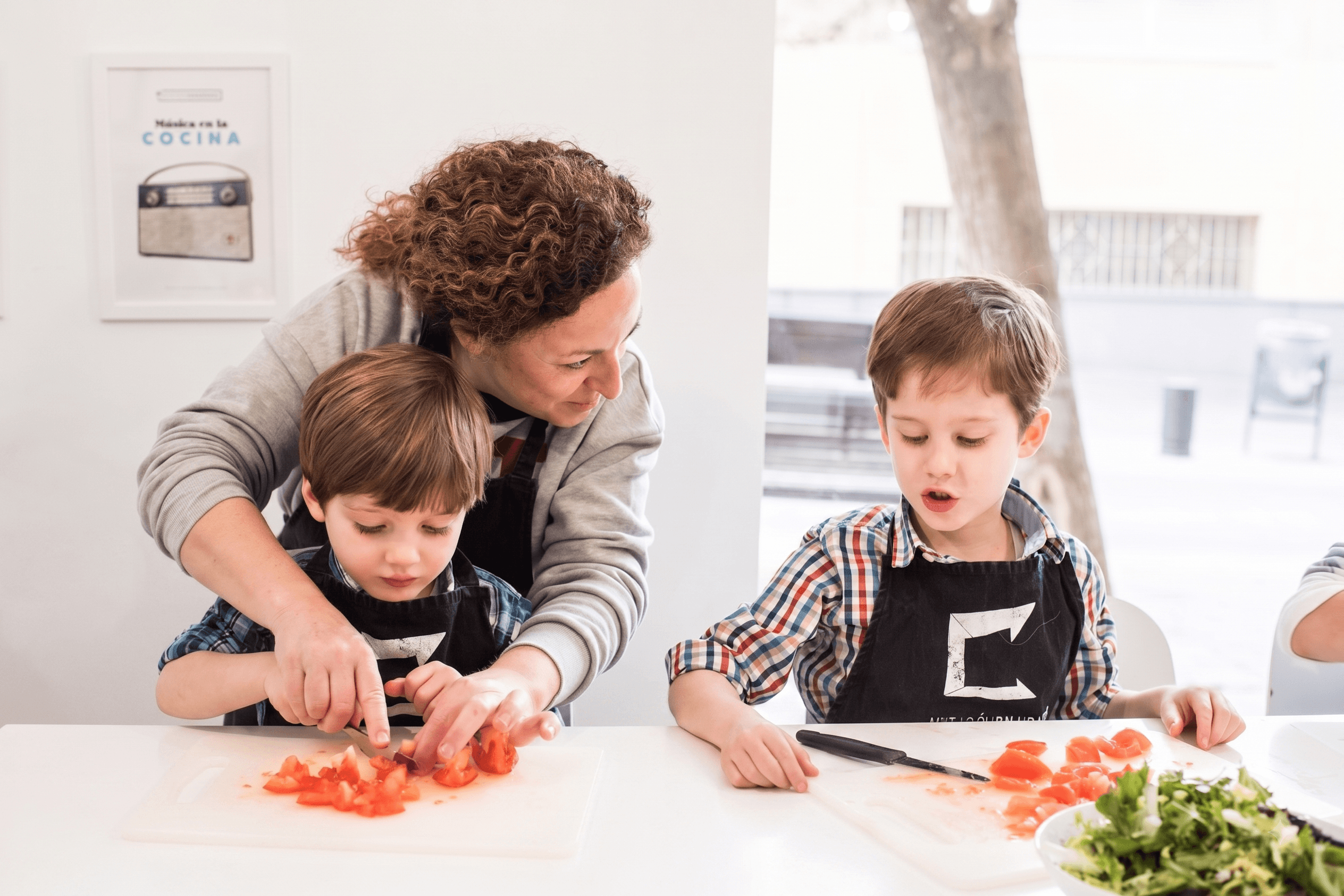 Una madre ayuda a su hijo a cortar tomate en un taller de cocina de La Habitación Saludable, junto a otro niño